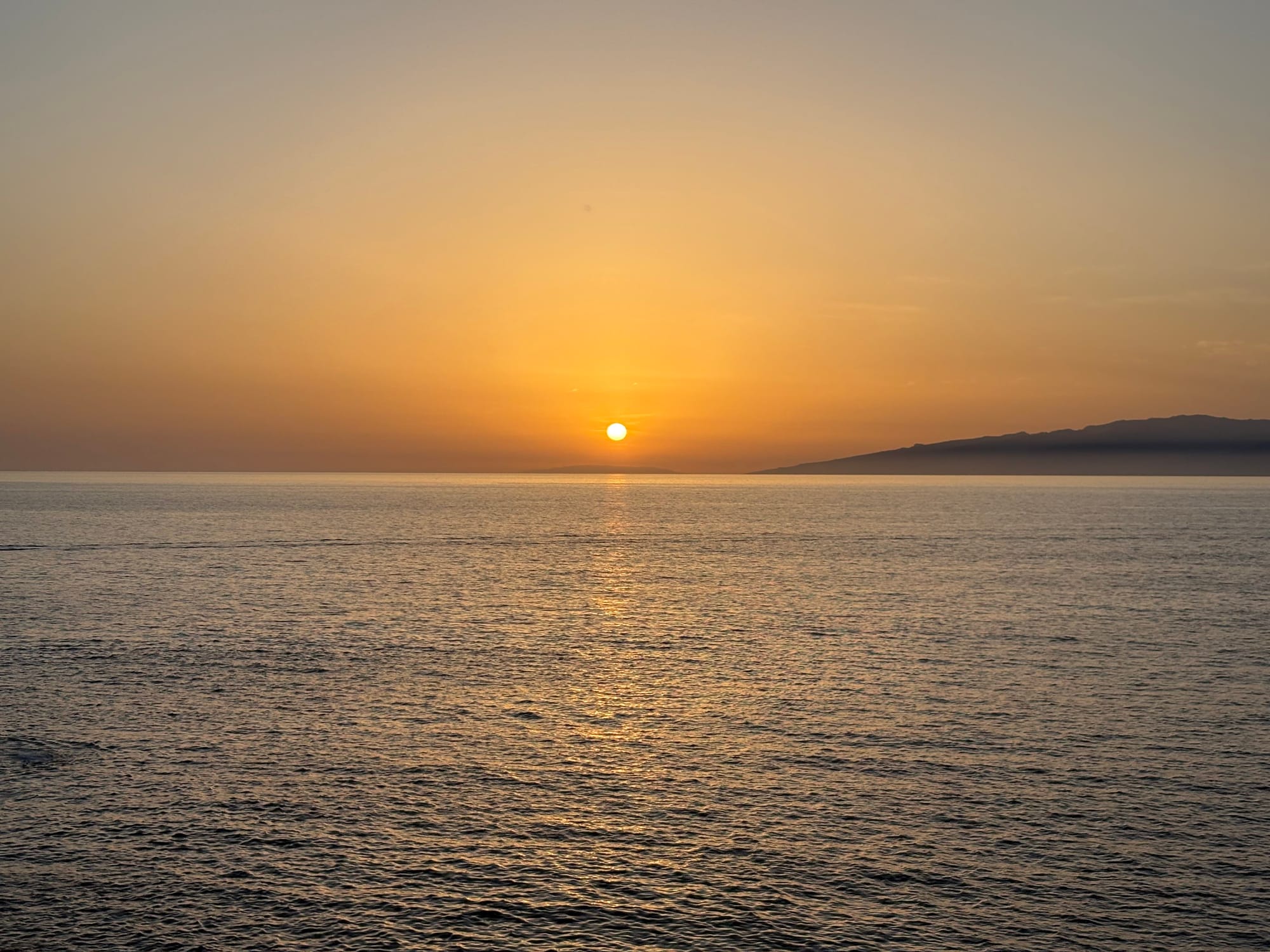 Golden sunset over calm sea, sun on the horizon, distant headland silhouetted on the right.